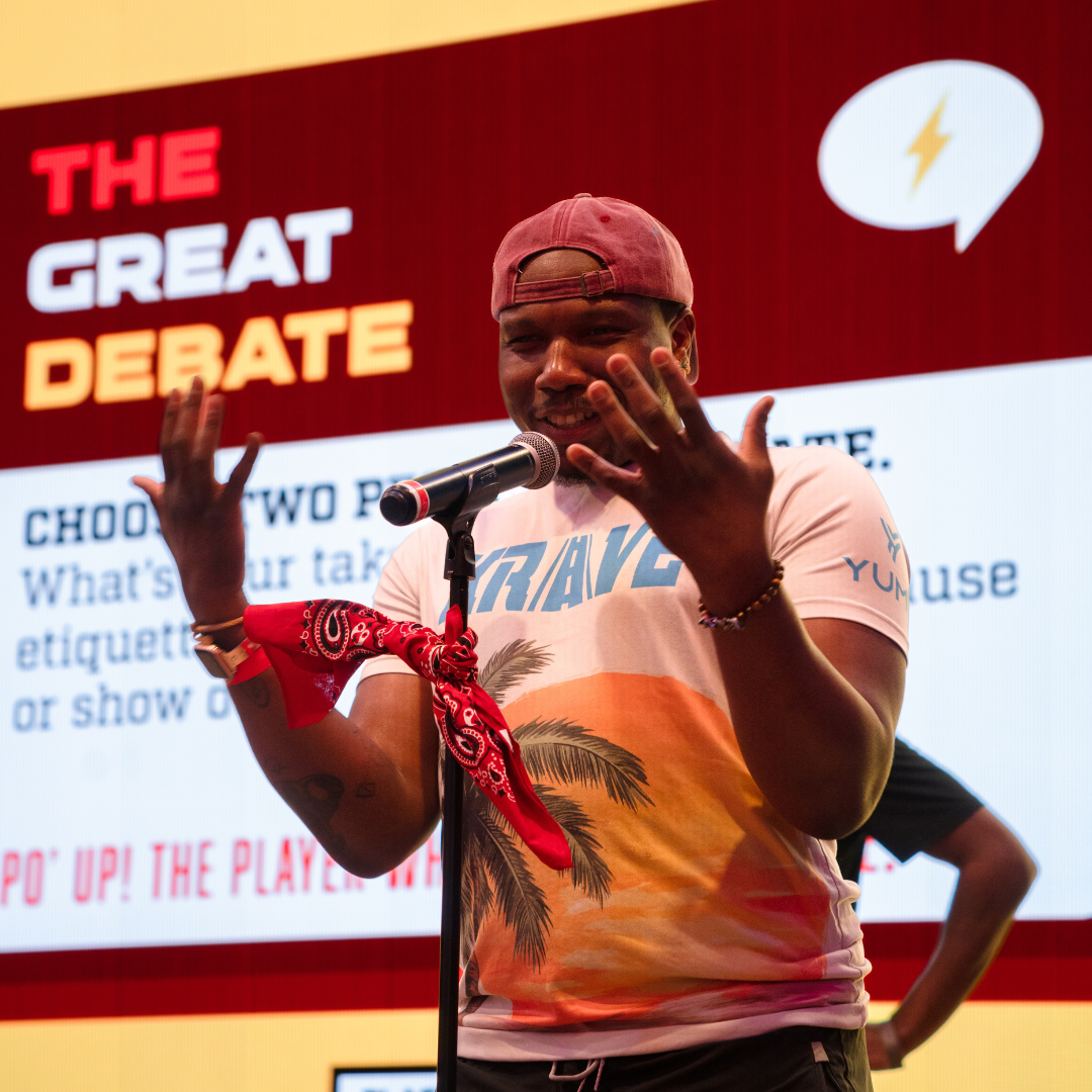 A Black man with a red baseball cap on stage playing PO' U! Live, answering a "Great Debate" question from PO' UP! Card Game: College Edition on graduation etiquette.