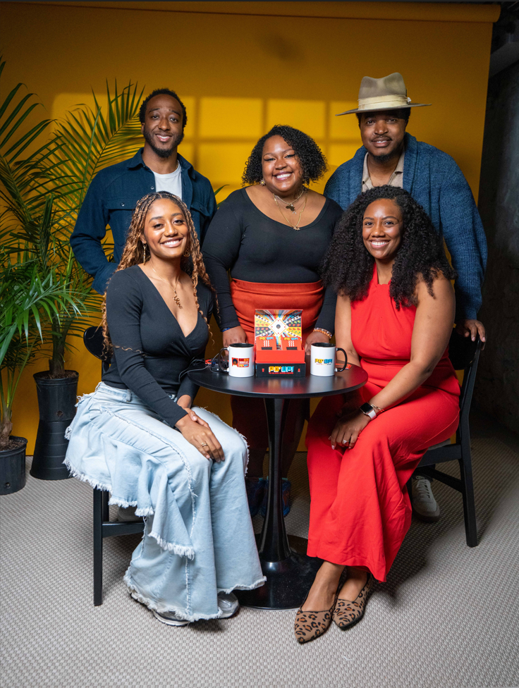 Group of five people posing together in a casual setting with a yellow wall and plants.